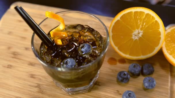 Αλκοόλ κοκτέιλ στο γυαλί βράχους με πορτοκάλι, πάγο και βατόμουρου στο τραπέζι, πλευρική θέα. — Αρχείο Βίντεο