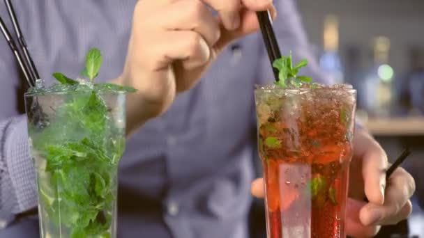 Barman bereidt alcohol cocktails met tequila, munt en ijs in de glazen. Zet rietjes. Close-up van de handen. — Stockvideo
