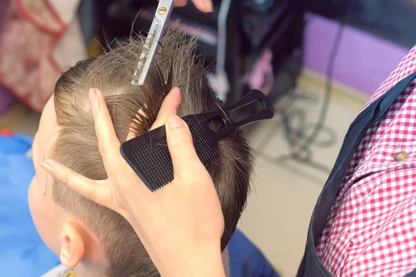 Hairdresser milling hairs with scissors on boys head. Top view, stylists hands close-up.