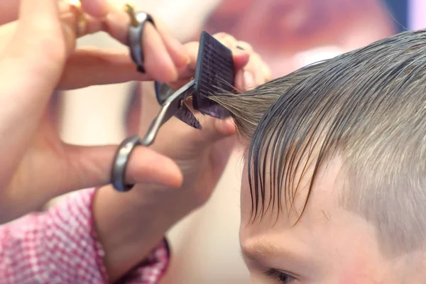Hairdresser cuts bangs with scissors on boys head. Stylists hands close-up, side view.