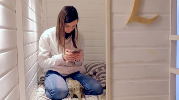Woman types a message on phone sitting in kids play house with meerkat. — Stock Video