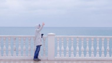 Kadın beyaz ceket ve kaput selfie sahildeki Deniz Manzaralı smartphone cep telefonu ile güzel terasta video yapar. Yan görünüm.