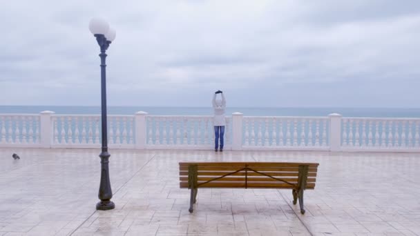 Frau in weißer Jacke und Kapuze auf wunderschöner Terrasse mit Meerblick am Wasser. zurück. — Stockvideo
