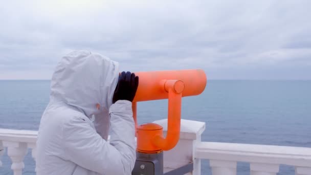 Žena v bílém plášti a kapucí vypadá v dalekohledu na moři nábřeží s výhledem na moře. Boční pohled. — Stock video