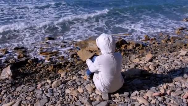 Woman in a white down jacket and hood sits on the shore and looks at the stormy sea waves beating against the stone shore. Back view. — Stock Video