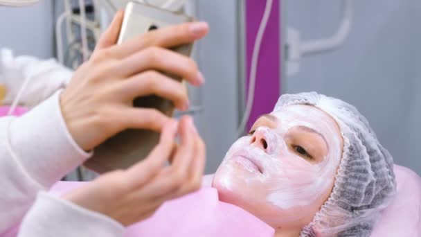 Mulher lê algo em um telefone celular deitado no procedimento no escritório de esteticistas com uma máscara no rosto . — Vídeo de Stock