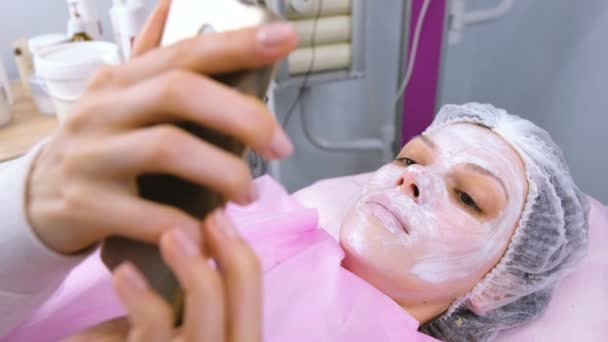 La mujer lee algo en un teléfono móvil tumbado en el procedimiento en la oficina de cosmetoloistas con una máscara en la cara . — Vídeos de Stock