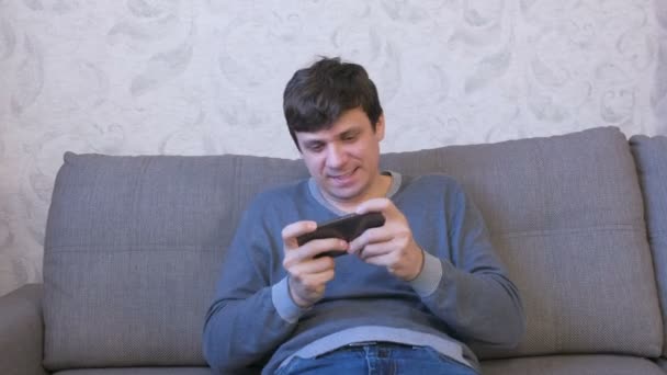幸せな男は、ソファーに座っていた彼の携帯電話でゲームをプレイします。. — ストック動画