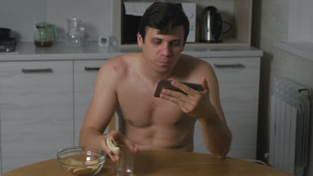 Νεαρός άνδρας τρώει μπισκότα στην κουζίνα το βράδυ και να αναζητούν βίντεο στο τηλέφωνο. Αϋπνία και το άγχος. Υπερβολικό φαγητό. — Αρχείο Βίντεο