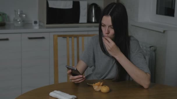 Mujer ve un vídeo divertido en el teléfono y se come una mandarina en la cocina por la noche . — Vídeos de Stock