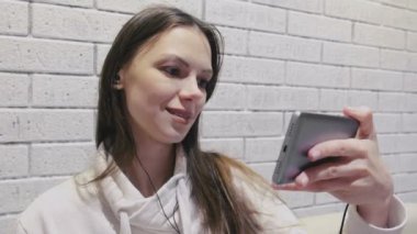Güzel esmer kadın romantik seria a seri halinde bir tuğla beyaz duvar arka planda cep telefonunda kulaklık, izlerken.