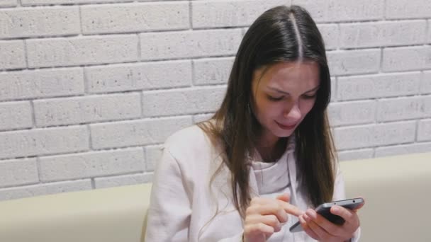 Mooie brunette lachende vrouw is te typen en het schuren van een bericht op mobiele telefoon zitten en wachten op iemand in café. — Stockvideo