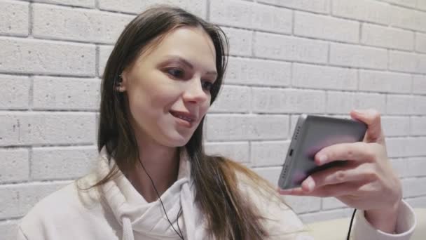 Mooie brunette vrouw kijken romantische seria voor een serial in koptelefoon op mobiele telefoon op een baksteen witte muur achtergrond. — Stockvideo