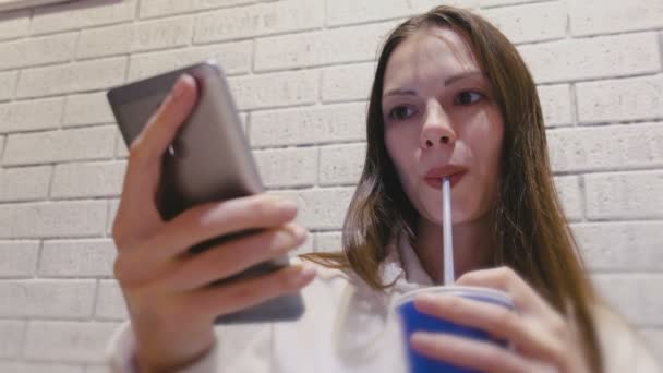 Sonriendo hermosa mujer está bebiendo una bebida de un tubo y viendo vídeo en el teléfono móvil en la cafetería . — Vídeos de Stock