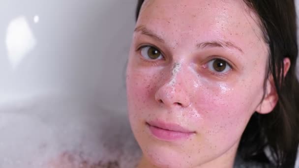 Hermosa mujer morena con exfoliante en la cara acostada en el baño en casa y sonriendo. Primer plano de la cara . — Vídeos de Stock