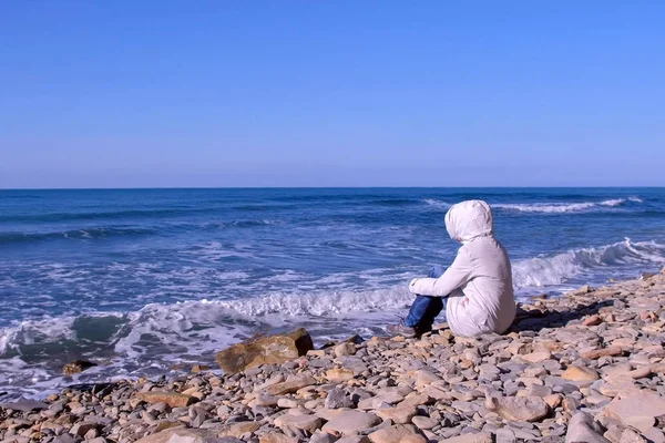 Mujer en una chaqueta blanca y capucha se sienta en la orilla y mira las tormentosas olas del mar golpeando contra la orilla de piedra . —  Fotos de Stock