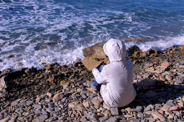 Femme dans une veste en duvet blanc et capuche se trouve sur le rivage et regarde les vagues de mer orageuses battant contre le rivage de pierre. Vue arrière . — Photo