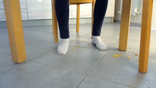 Frau isst am Tisch und zerbröselt Cornflakes auf dem Boden. Beine und Boden, Vorderansicht. — Stockvideo