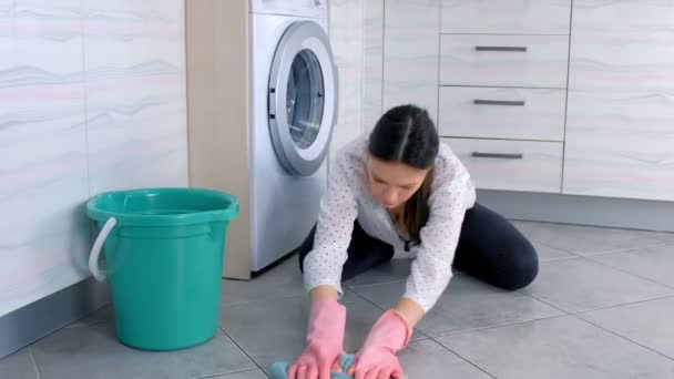 Mulher cansada em luvas de borracha rosa lava o chão da cozinha com um pano e olha para a câmera. Azulejos cinza no chão . — Vídeo de Stock