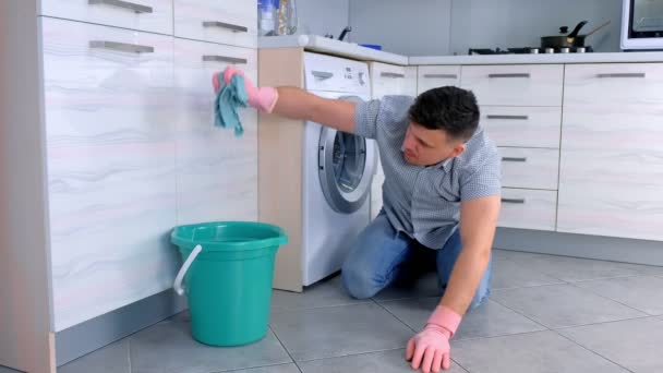Ευτυχισμένος άνθρωπος σε λαστιχένια γάντια πλένει και τρίβει σκληρά το πάτωμα στην κουζίνα, καθισμένος στο πάτωμα, πλευρική όψη. — Αρχείο Βίντεο