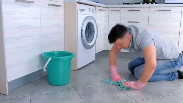 Unhappy tired man in rubber gloves washes and rubs hard the floor in the kitchen. Side view. — Stock Video