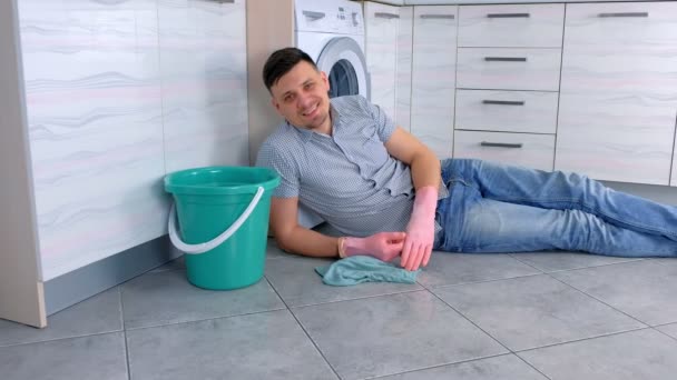 Heureux homme souriant dans des gants en caoutchouc a un repos de nettoyage pose sur le sol de la cuisine . — Video