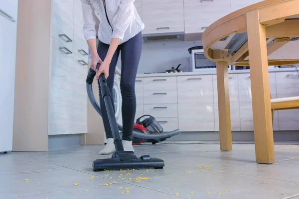 Woman is vacuuming the kitchen floor. She tidies up the corn flakes scattered on the gray tile.