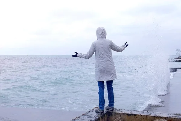 Mujer con una chaqueta blanca de pie en el paseo marítimo en la playa mira las olas de tormenta. Vista trasera . —  Fotos de Stock