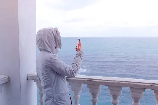 Mujer en la chaqueta blanca dispara vídeo olas del mar en el teléfono inteligente en la hermosa terraza con vista al mar. Vista trasera . —  Fotos de Stock