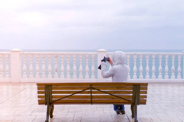 白いジャケットとフードの女性は、海の波ウォーター フロントの海を望む美しいテラスのベンチに座ってのスマート フォンでビデオを撮影します。背面図. — ストック写真