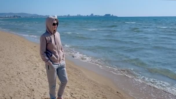 Žena v tričku a slunečních brýlích stojí na písečné pláži za jasného slunečného dne.