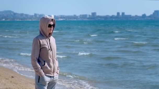 Kobieta w bluzę i okulary przeciwsłoneczne stoi na piaszczystej plaży na jasnym słoneczny dzień. — Wideo stockowe