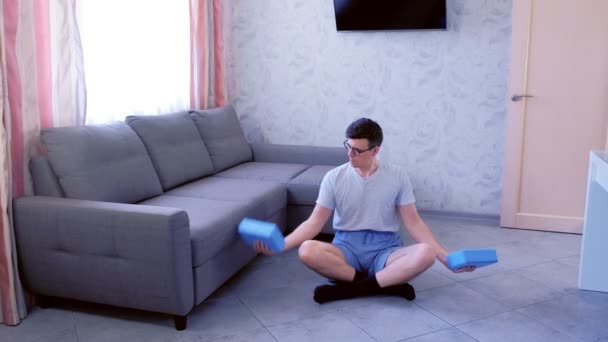 Funny Nerd man gör övningar för hand biceps med yoga block i stället för hantlar med insatser sitter på golvet hemma. Sport humor koncept. — Stockvideo