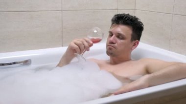 Sarhoş yetişkin adam köpük banyosunda kırmızı şarap içiyor.