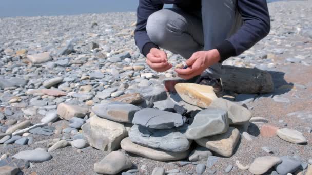 Człowiek zapala się węglem w ogniu z meczów na kamiennej plaży, ręce zbliżenie. — Wideo stockowe