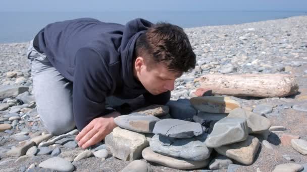 Чоловік дме вугілля в багатті на кам'яному морському пляжі. Створення вогнища кампусу . — стокове відео