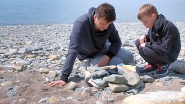 Otec a syn zapalují uhlí v ohni na ručně vyráběné kameny a mluví na mořské pláži. — Stock video