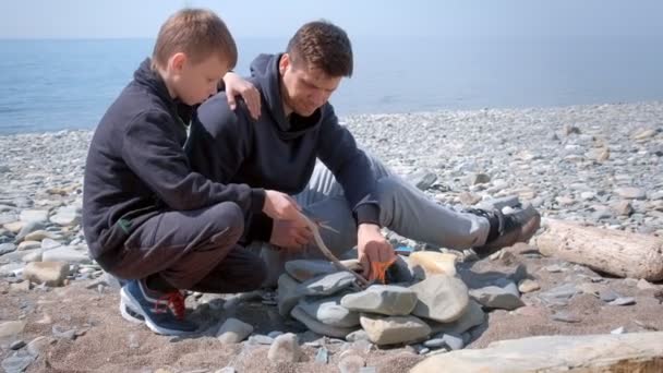 Baba ve oğlu deniz sahilinde ki el yapımı taşların üzerindeki şenlik ateşindeki kömürleri tutuşduruyorlar. Aile kamp. — Stok video