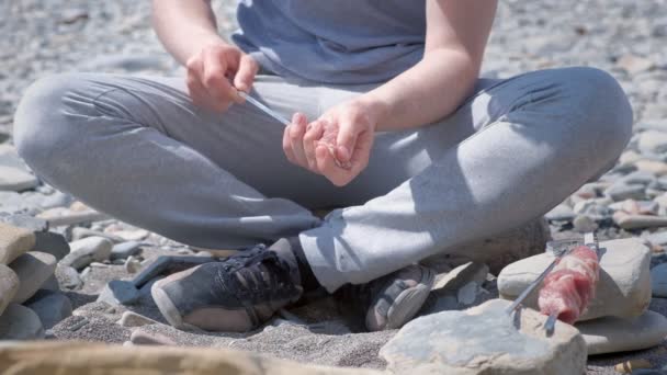 Man is preparing shashlik putting the slices of meat on the skewer sitting on stones, hands closeup. — Stock Video