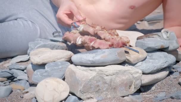 Man is koken shashlik op kampvuur roterende spiesjes liggend op de stenen, voedsel close-up. — Stockvideo
