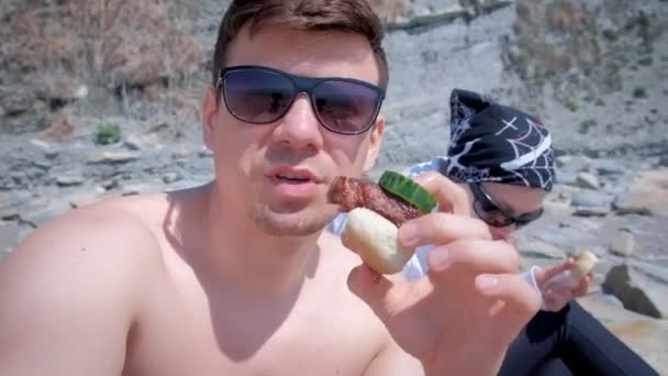 Man blogger is eating shashlik with bread and cucumber on camera with his wife and talking about it his subscribers. — Stock Video