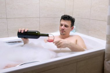 Yorgun sarhoş adam banyoda yatıyor, bir şarap bardağına şarap dökün ve içecek.