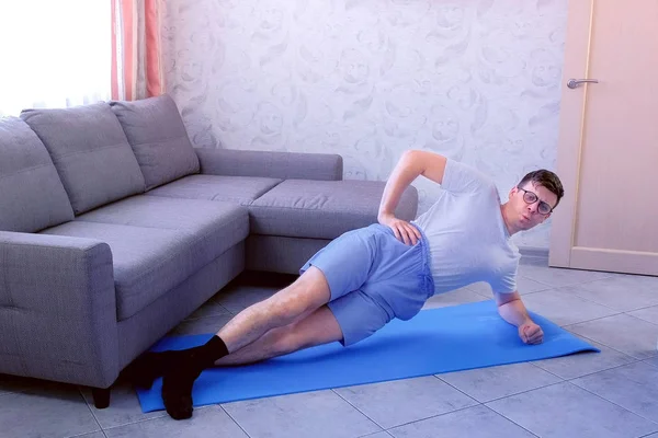 Zwakke grappige nerd man maakt dynamische kant plank met inspanningen op de mat thuis. — Stockfoto