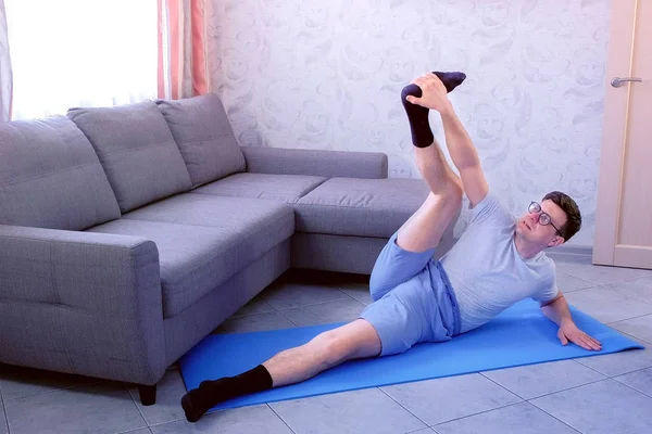 Crazy nerd man is trying to stretch his legs laying on the mat at home. Sport humor concept. — Stock Photo, Image
