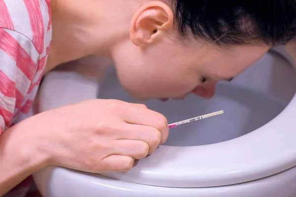 Morning sickness. Young pregnant tired woman with pregnancy test in hand is vomiting in toilet at home, closeup view.