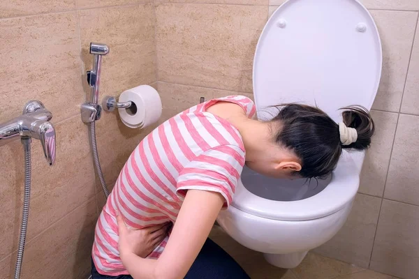 Chory młoda kobieta jest wymioty w toalecie siedzi na podłodze w domu, objawem zatrucia pokarmowego. — Zdjęcie stockowe