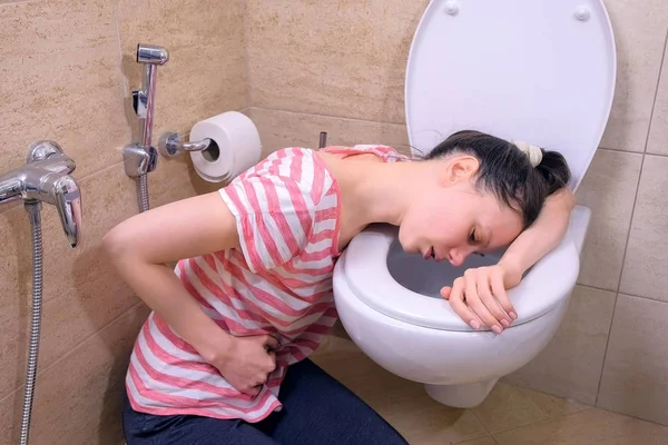 Zieke jonge vrouw met pijn in de maag is braken in toilet zittend op de vloer thuis, voedselvergiftiging symptoom. — Stockfoto
