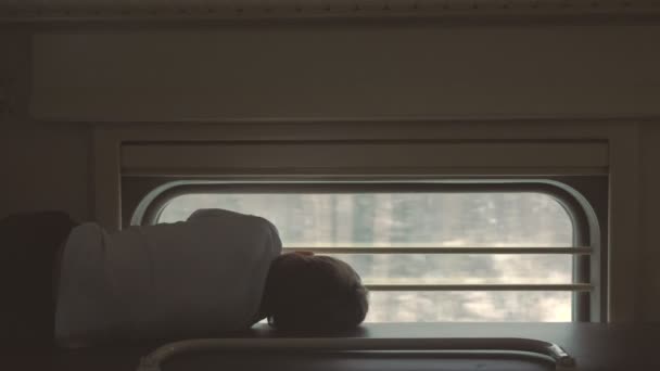 Jongen op de trein liggend op de bovenste plank in een tweederangs koets en venster te kijken. Achteraanzicht. — Stockvideo