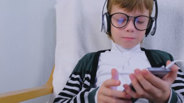 Niño en gafas está escuchando música en los auriculares en el teléfono inteligente sentado en el sillón en casa . — Vídeos de Stock
