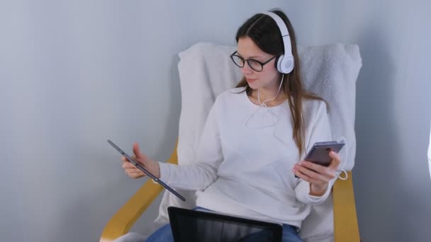 Woman in glasses and headphones is working on laptop sitting in armchair. Mobile phone and tablet in hands. — Stock Video